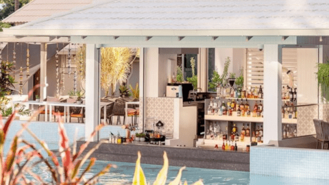 A poolside bar at the Novotel Cairns Oasis Resort features a variety of bottles on shelves, a white roof, and a seating area surrounded by lush plants.