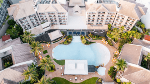 Aerial view of Novotel's hotel complex in Cairns, featuring a large pool surrounded by lounge chairs, palm trees, and landscaped gardens, with multi-story buildings in the background.