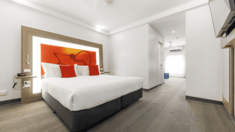 Located in the heart of Darwin's CBD, this modern Novotel hotel room boasts a large bed with white linens and red accent pillows. The brightly lit headboard complements the white walls, and an open doorway leads to a well-lit hallway.