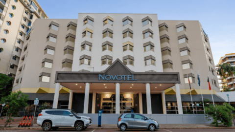 Front view of a multi-story Novotel building in the CBD of Darwin, featuring a vehicle parking area with two cars parked in front. The entrance boasts a covered porch supported by multiple pillars, and flags are displayed on the right side.