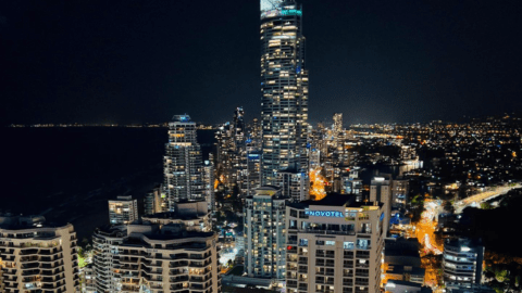 Nighttime cityscape of Surfers Paradise featuring illuminated high-rise buildings, with the tallest building, Novotel, in the center. City lights are visible in the background, creating a vibrant urban glow.
