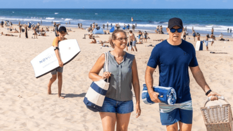A couple walking on a crowded beach near the Novotel, carrying beach gear, with a beachgoer holding a bodyboard in the background. The vibrant energy of Surfers Paradise makes it an ideal destination for sun and surf enthusiasts.