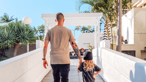 A tattooed man and a child holding hands walk down a sunlit pathway in Surfers Paradise, lined with palm trees and white structures, their destination the welcoming entrance of Novotel.