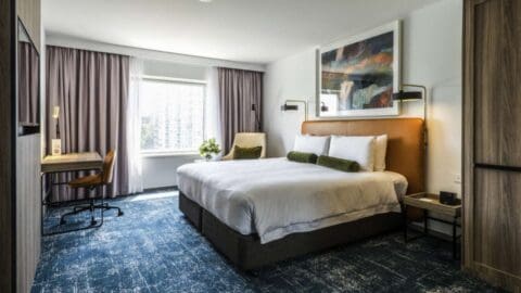 A modern hotel room at Novotel Sydney with a large bed, bedside lamps, a desk, a chair, and a window with curtains. The room features a neutral color scheme and a large abstract painting above the bed. Located near Darling Square, it's perfect for both business and leisure travelers.