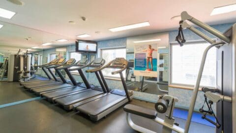 A gym at Novotel Darling Square in Sydney features treadmills lined up against a mirrored wall, various exercise machines, and motivational fitness posters. The space is brightly lit with overhead lights and large windows letting in natural light.