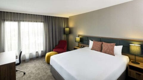 A Novotel Sydney room with a large bed, nightstands with lamps, a desk and chair, and an armchair near a window boasting sheer and blackout curtains. This Airport hotel offers modern decor in neutral tones for travelers at Sydney International Airport.
