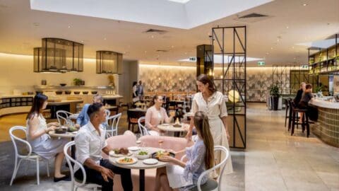 A modern restaurant at Novotel Sydney International Airport with patrons dining at tables, engaged in conversation. A waitress stands near a table. The interior features stylish decor and ample lighting.
