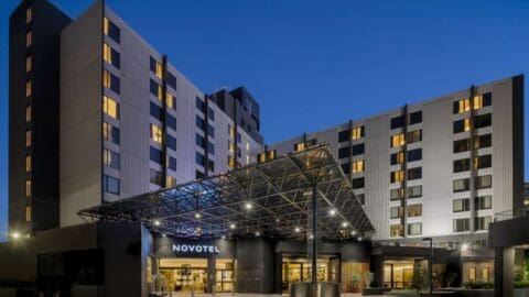 The image shows a modern multi-story Novotel hotel building at dusk, prominently displaying its name above the entrance, which is covered by a large glass and metal canopy. This stylish accommodation is conveniently located near Sydney's International Airport.