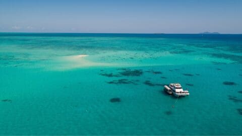 Aerial view of a turquoise ocean with a small sandy island and a yacht anchored nearby under a clear blue sky, perfect for Cairns Reef Tours.