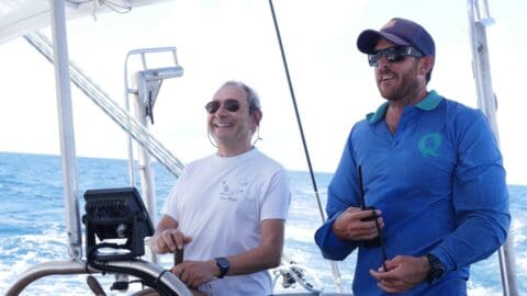 Two men steering a boat on the ocean, one wearing a white t-shirt with sunglasses, the other in a blue shirt wearing a cap and sunglasses. They are guiding an island tour near Cairns, exploring its stunning reef.