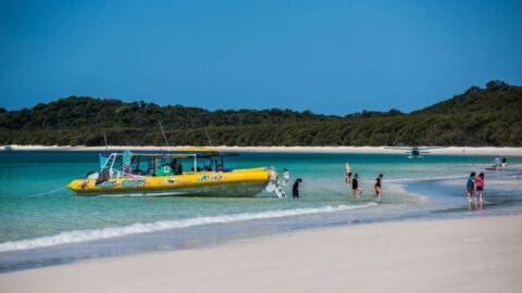 A group of people are disembarking from a yellow tour boat onto a sandy beach, with lush hills in the background and a clear blue sky overhead.