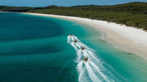 Aerial view of a white sandy beach with turquoise water. Several speedboats with yellow tops create white wakes as they travel parallel to the shore. The coastline is lined with sustainable, lush vegetation, promoting ecotourism.