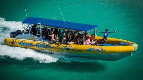 A brightly colored tour boat speeds across turquoise waters, carrying a group of smiling passengers and a guide in a blue shirt with arms outstretched. The boat, labeled "Ocean Rafting," provides an exciting and sustainable adventure on the water.