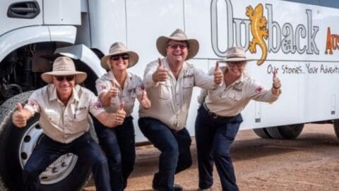 Four people wearing cowboy hats and beige shirts, posing with thumbs up in front of a vehicle with "Outback Tour" written on it.