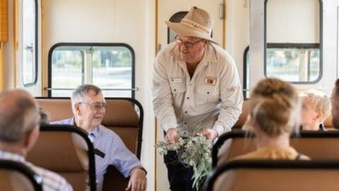 A guide in an outback hat and shirt shows native plants to passengers inside a sustainable ecotourism train.