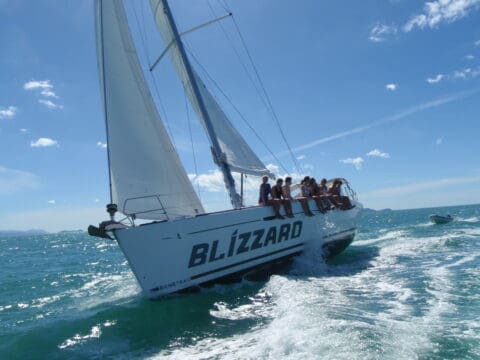 A white sailboat named "BLIZZARD" glides through the tranquil waters of the Whitsundays, with several people relaxing on the deck. The ocean is calm, and the sky is clear with a few clouds.