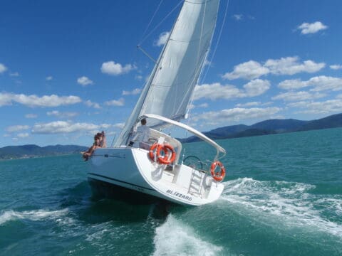 A sailboat named "Blizzard" elegantly navigates the clear blue waters of the Whitsundays, with two people on board under a sunny sky dotted with scattered clouds and distant green hills in the background.