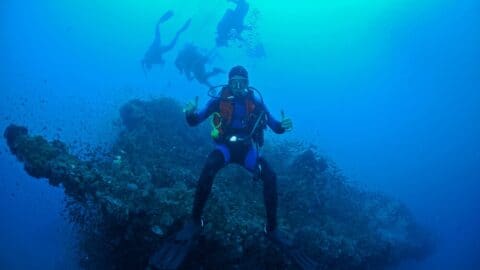 A scuba diver gives two thumbs up near the coral reef of Yongala, while other divers enjoy the dive in the background.
