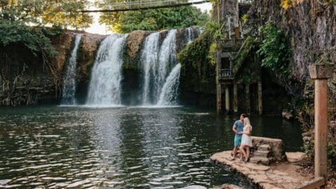 A couple stands on a stone platform beside a pool with a waterfall in the background, surrounded by lush vegetation and standing structures, highlighting an ecotourism adventure.