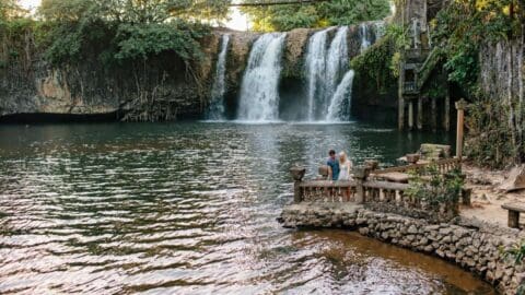 Two people stand by the water on a stone structure in front of a waterfall surrounded by lush nature, enjoying their tour.