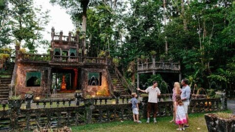 A certified tour guide is explaining the history of an ancient, ornate stone structure surrounded by lush trees to a family with two children in a park setting, emphasizing the principles of ecotourism.