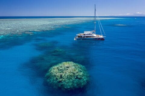 A sailing catamaran floats on clear blue water above a coral reef, with a wide expanse of the Great Barrier Reef visible in the background under a blue sky. This sustainable tour offers an exceptional ecotourism experience.