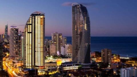 Nighttime cityscape featuring illuminated high-rise buildings in Broadbeach near a coastline, with a clear sky and urban lights visible in the foreground.