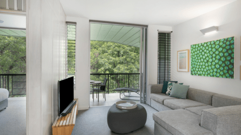 A modern living room with a gray sofa, round ottoman, wall art, TV, and sliding glass doors opening to a balcony with table and chairs overlooking greenery at Peppers Noosa Resort. The adjoining bedroom on the left adds to the villa's charm.