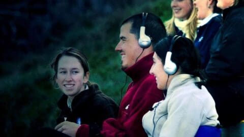 A group of people seated outdoors, some wearing headphones and smiling. They are dressed warmly, suggesting a cool environment, indicative of a sustainable approach to enjoying nature.