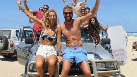 A group of smiling friends pose on and around a pair of parked off-road vehicles on a sunny beach, raising their arms in celebration during their ecotourism tour.