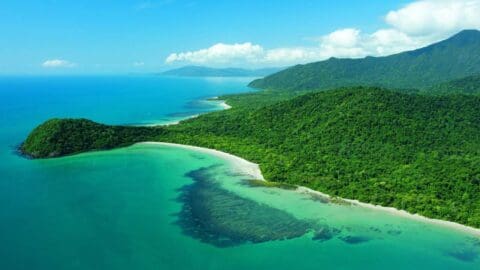 Aerial view of a lush green coastline with clear blue ocean waters and sandy beaches under a slightly cloudy sky. Dense forests cover the hills and mountains in the background.