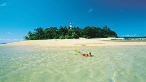 A person snorkeling in clear, shallow waters near a sandy beach with an island and a lighthouse in the background.