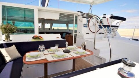 The image shows the deck of a yacht with a dining area set for four, including plates, glasses, and flowers. In the background, there are navigational controls and seating. The clear and sunny sky enhances the beautiful connection to nature.