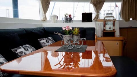 A polished wooden table with a decorative metal carriage holding three small plants sits in a room with black cushioned seating and patterned pillows, creating a cozy nature-inspired atmosphere. A counter with drinkware and decor is in the background.