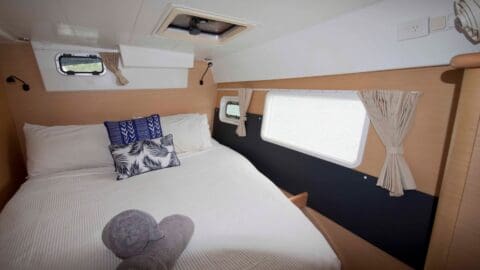 A compact camper van interior featuring a neatly made bed with white linens, two patterned pillows, rolled towels on the bed, and windows with beige curtains on the side and rear walls offers a cozy and sustainable accommodation close to nature.