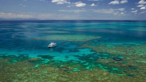 A tour boat floats on clear, turquoise waters above a diverse coral reef system under a partly cloudy sky.