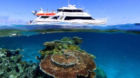 A tour boat named Poseidon floats on the clear blue ocean surface, above colorful coral formations underwater. Four people are visible on the boat's upper deck.