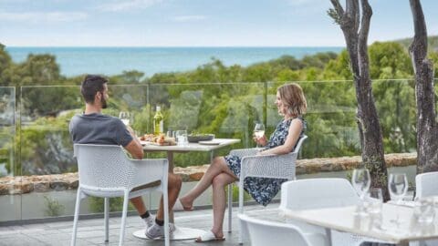 A couple sitting at a decked table on a terrace in front of green luscious hills and the blue ocean in the far.