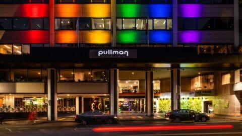 A hotel facade lit up with rainbow colors at night, featuring the Pullman Hotels sign above the entrance. Cars with light trails can be seen on the street in front, adding vibrancy to the scene near Hyde Park in Sydney.
