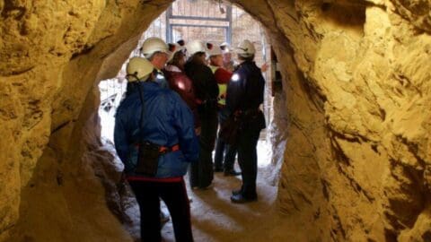 A group of people wearing helmets and safety gear stands inside a well-lit tunnel with rocky walls, participating in an ecotourism adventure.