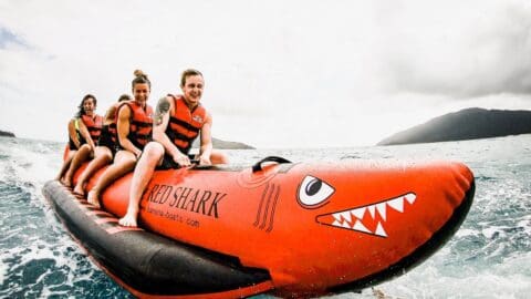 Three people wearing life vests ride a red inflatable banana boat named "Red Shark" on the water, with a cloudy sky and mountains in the background. This thrilling ride is part of the exciting Red Cat Adventures experience.