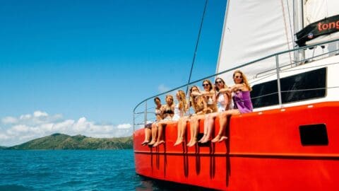 A group of people sits on the edge of a red boat from Red Cat Adventures, sailing on clear blue water under a sunny sky, with a green hilly landscape in the background.