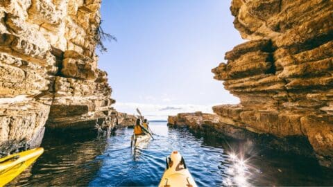 A person in a kayak navigates a narrow waterway between two rocky cliffs under a clear blue sky, immersing themselves in the sustainable beauty of nature.