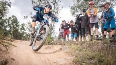 A mountain biker wearing a helmet rides down a dirt trail while a group of spectators watches and cheers in the background. Trees and vegetation surround the area, showcasing an ecotourism spot supporting sustainable outdoor activities.