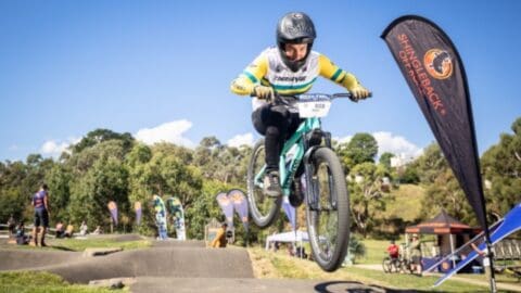 A cyclist wearing a helmet and colorful jersey is airborne while riding a mountain bike over a dirt track. Trees and banners are visible in the background, highlighting the excitement of this sustainable tour through nature.