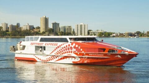 A red and white Rottnest Express ferry, promoting ecotourism, sails on calm water with city buildings visible in the background.