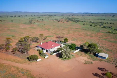 Aerial view of a rural property with a red-roofed house, surrounded by sparse trees and bordered by open farmland under a clear blue sky, reminiscent of the serene landscapes found in the Bendleby Ranges.