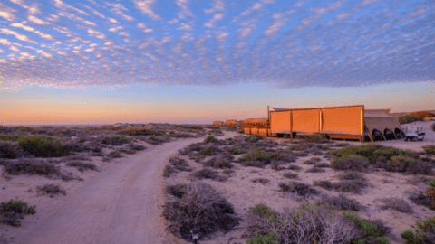 A sandy path leads to a small settlement with several tent-like structures at dusk, under a sky full of scattered clouds. Sparse vegetation dots the desert landscape, hinting at the rugged beauty near Sal Salis and the pristine Ningaloo Reef.