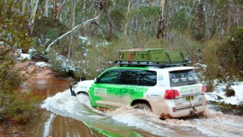 A green and white off-road SUV from Simmo's Tours drives through a muddy, shallow water crossing in a forested area with snow on the ground. The vehicle has a roof rack carrying various equipment, showcasing their expertise in offroad adventures.