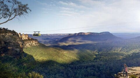 A couple stands at a scenic viewpoint overlooking a vast valley with rugged cliffs, while a cable car descends on the left side, offering an immersive nature tour.